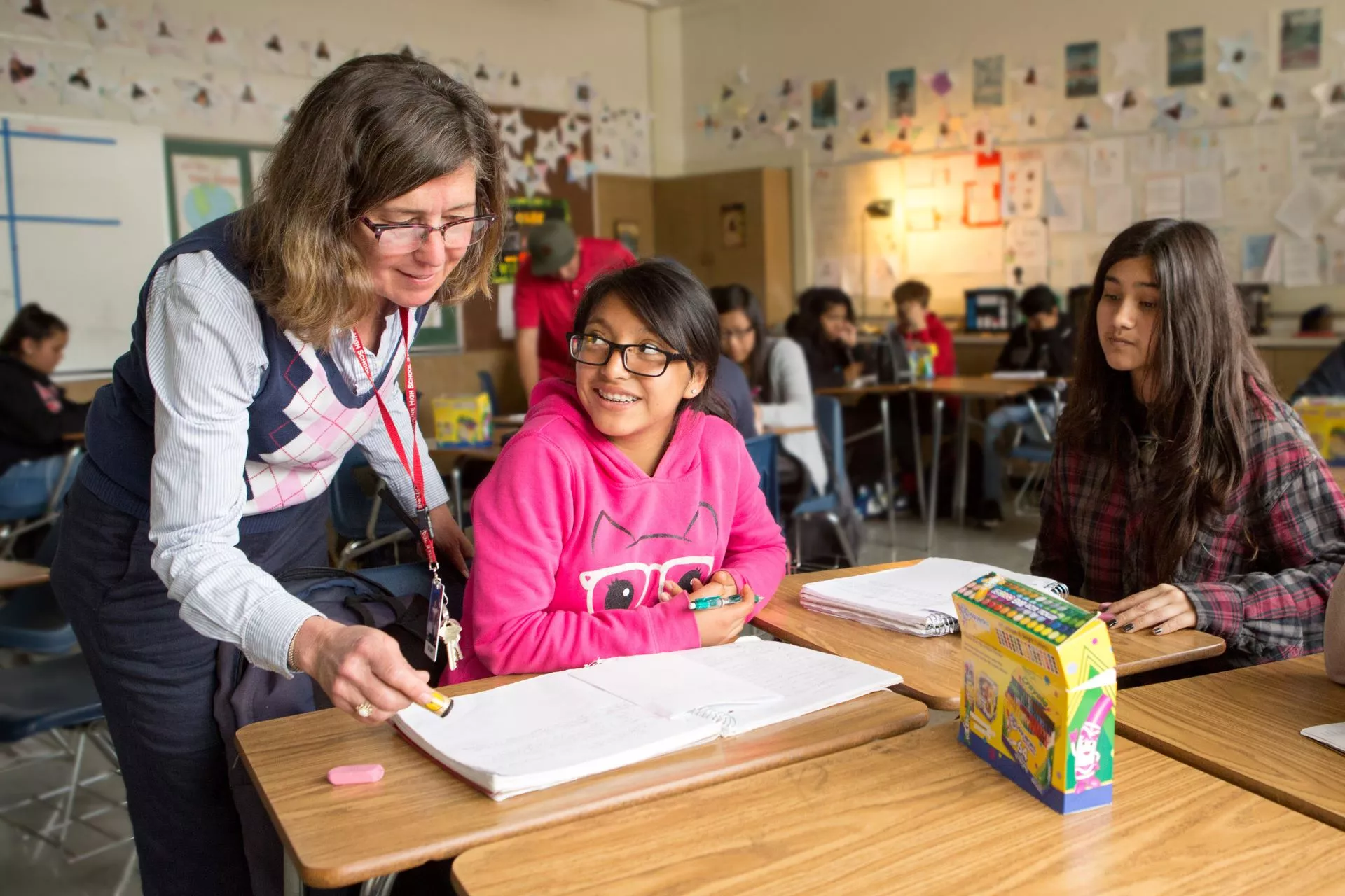 In a middle school classroom a female educator leans over a desk where two students are sitting and inspects their work