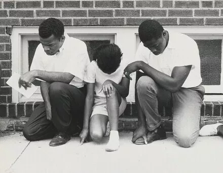 Three young students of color kneel in prayer in front or a brick wall.