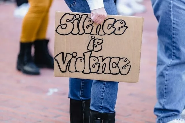 Someone holds a cardboard sign to their side. The sign reads, in large bubble letters: "Silence is violence."