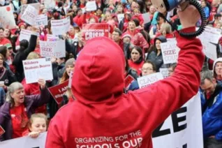 A person stands holding a blue megaphone with their back to the camera in a red hoodie with the hood up. In front of them there is a huge crowd of people dressed in Red for Ed.