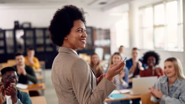 A teacher stands in front of her classroom. She looks back over her shoulder towards the camera, pointing with a pen.