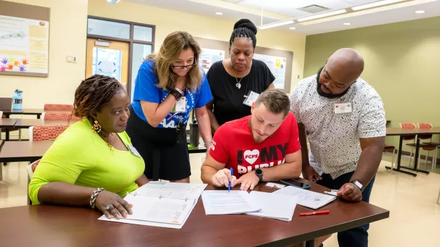A group of 5 educators gather around a table in a classroom, reviewing language in a contract.