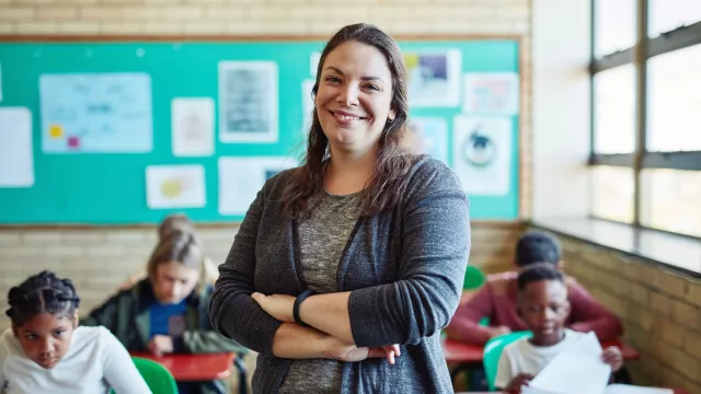 a female teacher stands in front of a classroom, she smiles with her arms crossed.