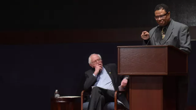 a black man at a lecturn with Sen. Bernie Sanders looking on