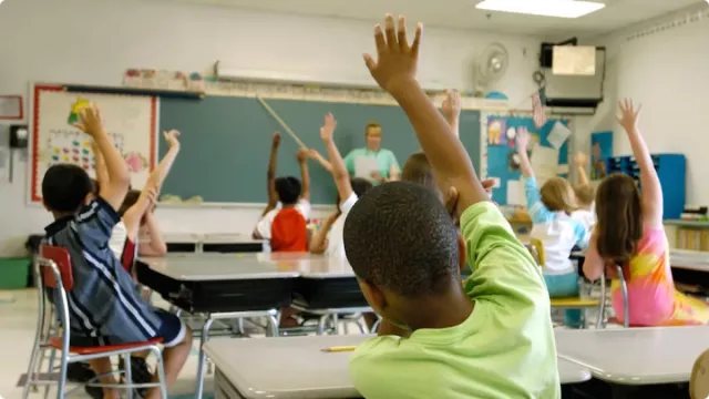 A diverse classroom of students faces a blackboard and raises their hands