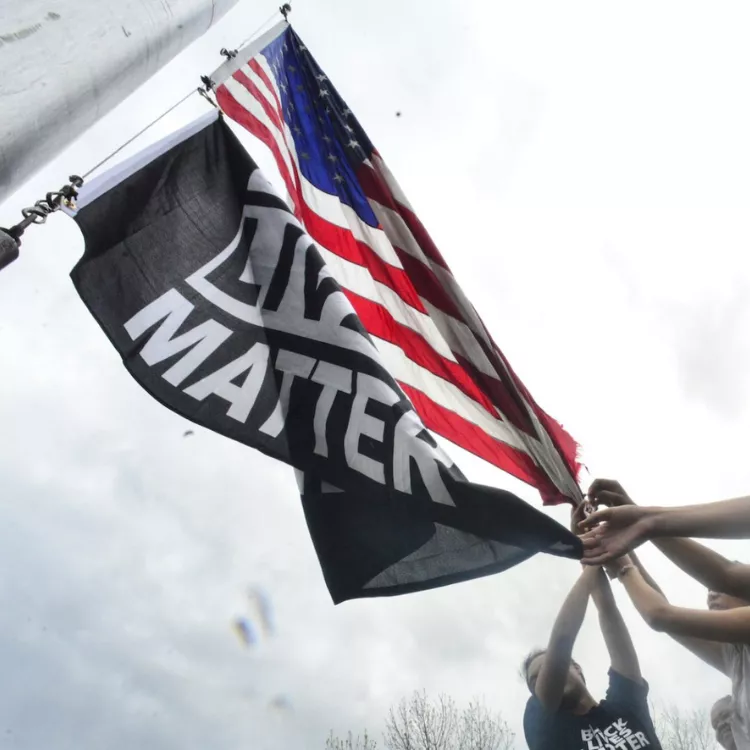 An American flag and a Black Lives Matter flag are raised on a flag pole by several pairs of hands. The hands visible include people of multiple races. 