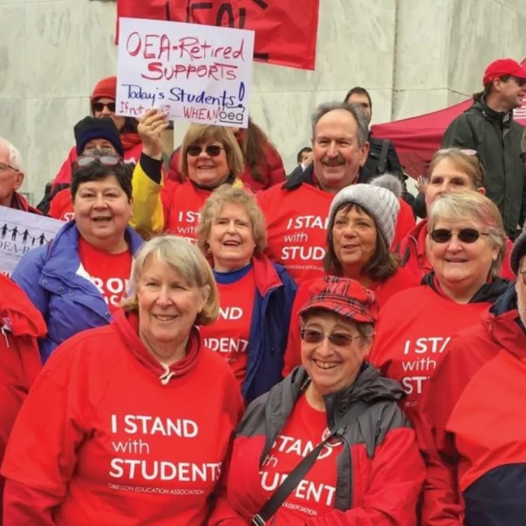 A group of OEA-Retired members stand smiling together at a rally. They are all dressed in red, with many shirts that say, "I stand with students." In the background, one OEA-Retired Member holds a sign that says, "OEA-Retired supports Today's students. If not now...when?!"